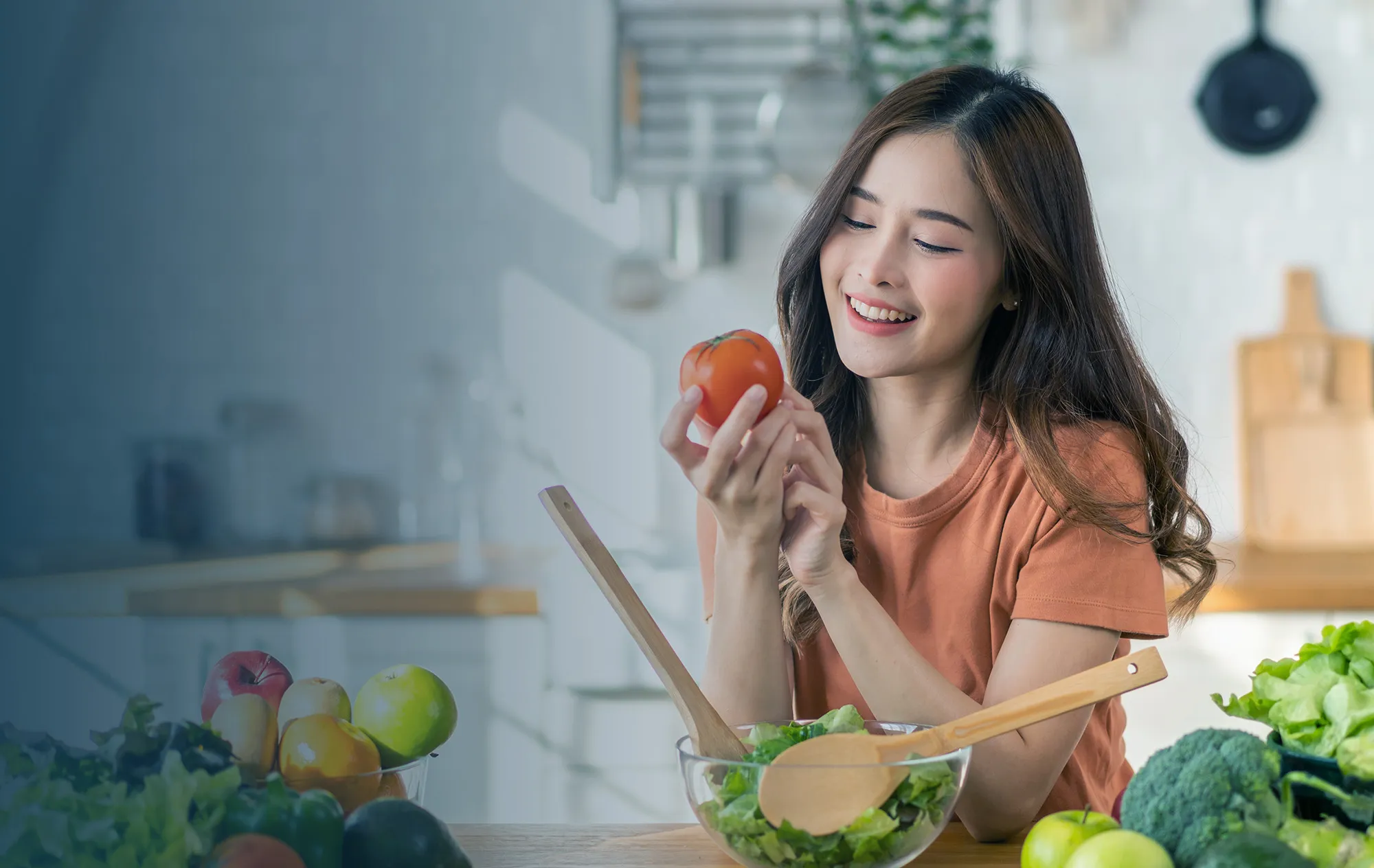 A smiling woman holding a tomato at a kitchen counter, with a bowl of salad, fresh fruit and vegetables in front of her