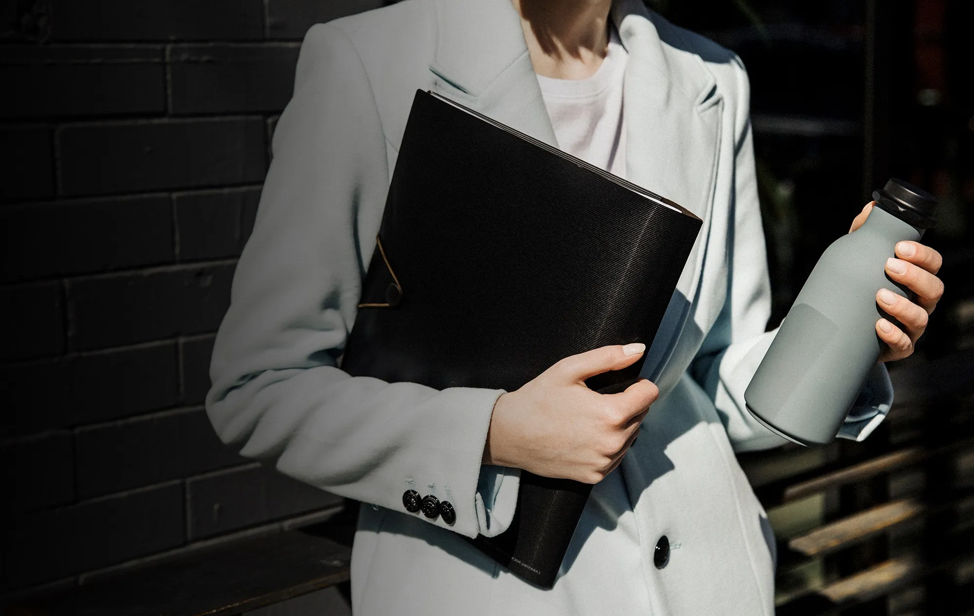 A woman in a pale blue coat holding a black folder against her side and a grey-green water bottle in her other hand, beside a black brick wall