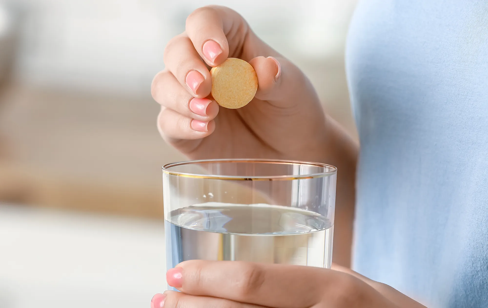 A hand with pink nails holding a round pale orange tablet above a glass of water held in the other hand
