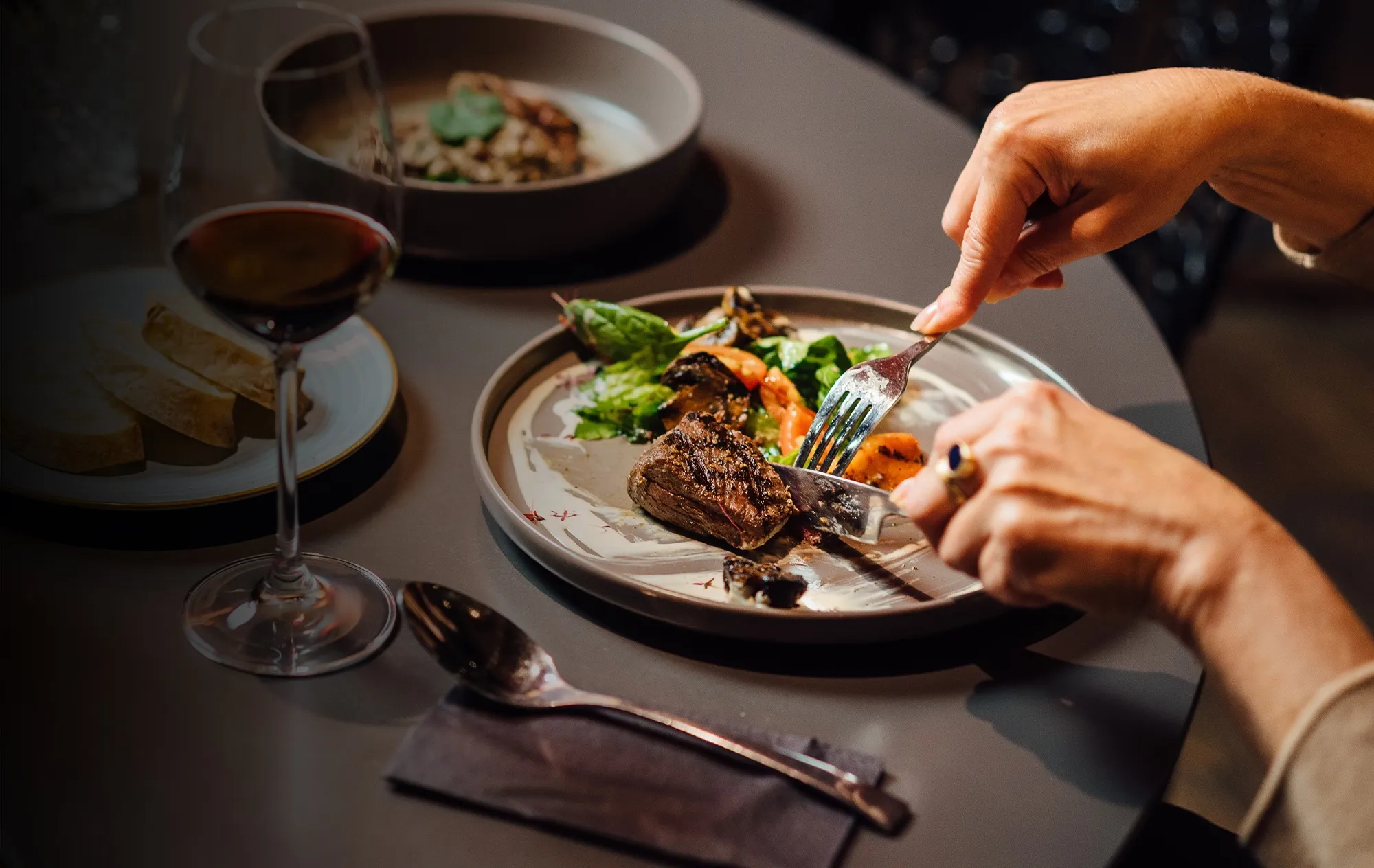 A diner cutting a grilled steak with salad on a grey plate, with a glass of red wine and a plate of bread on the table