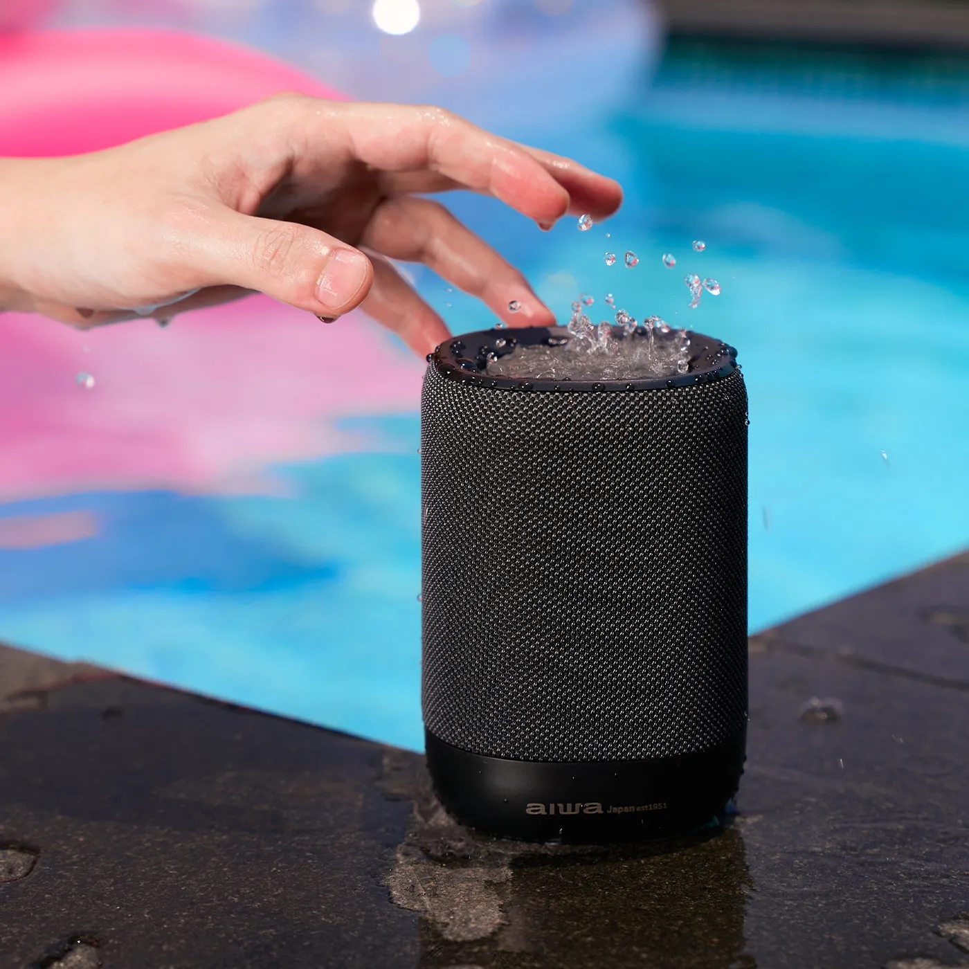 A wet hand above a black AIWA speaker splashed with water on the stone edge of a blue swimming pool, a pink float behind