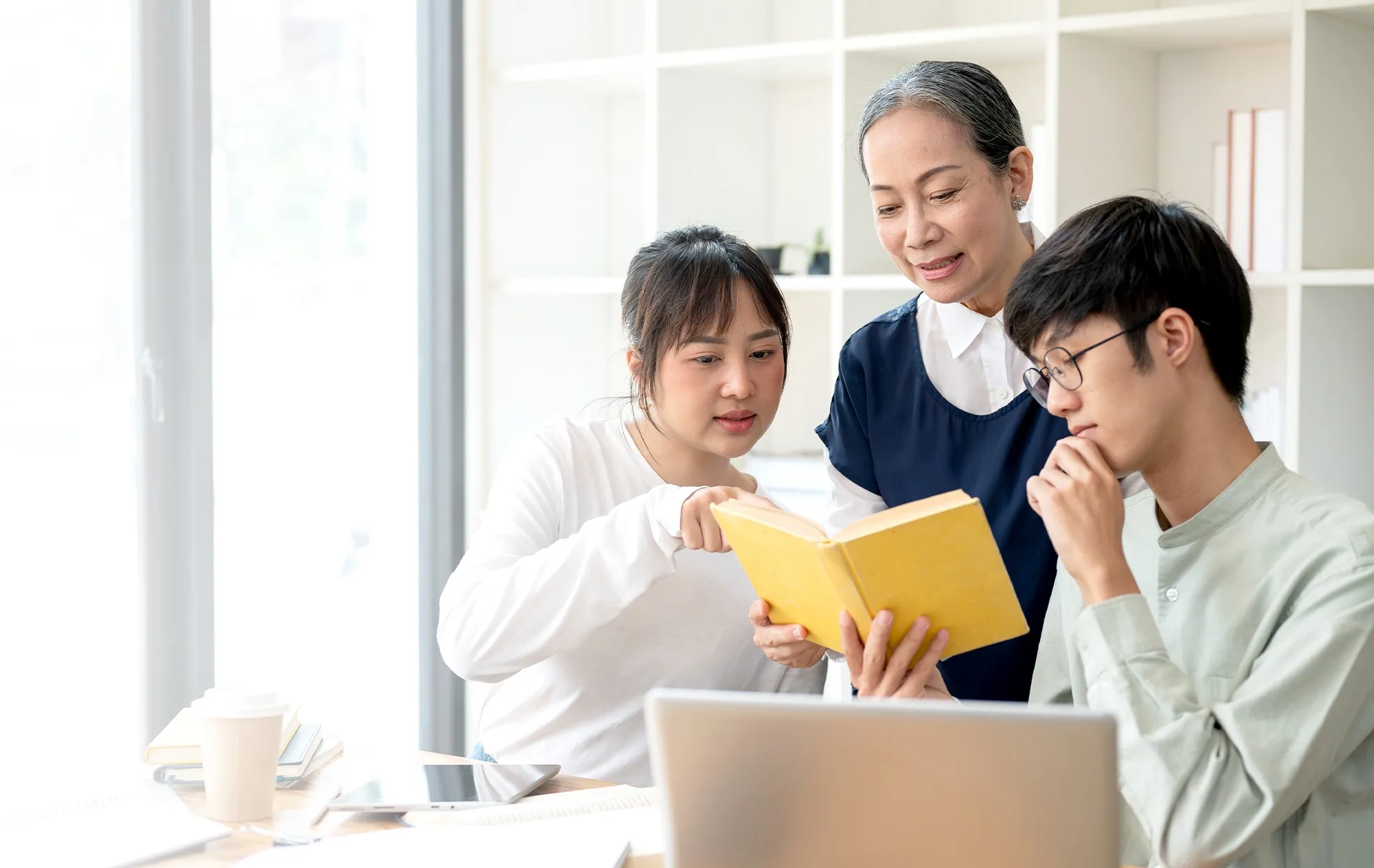 A young woman pointing at a page of an open yellow book as an older woman and a young man in glasses read along at a desk with a laptop