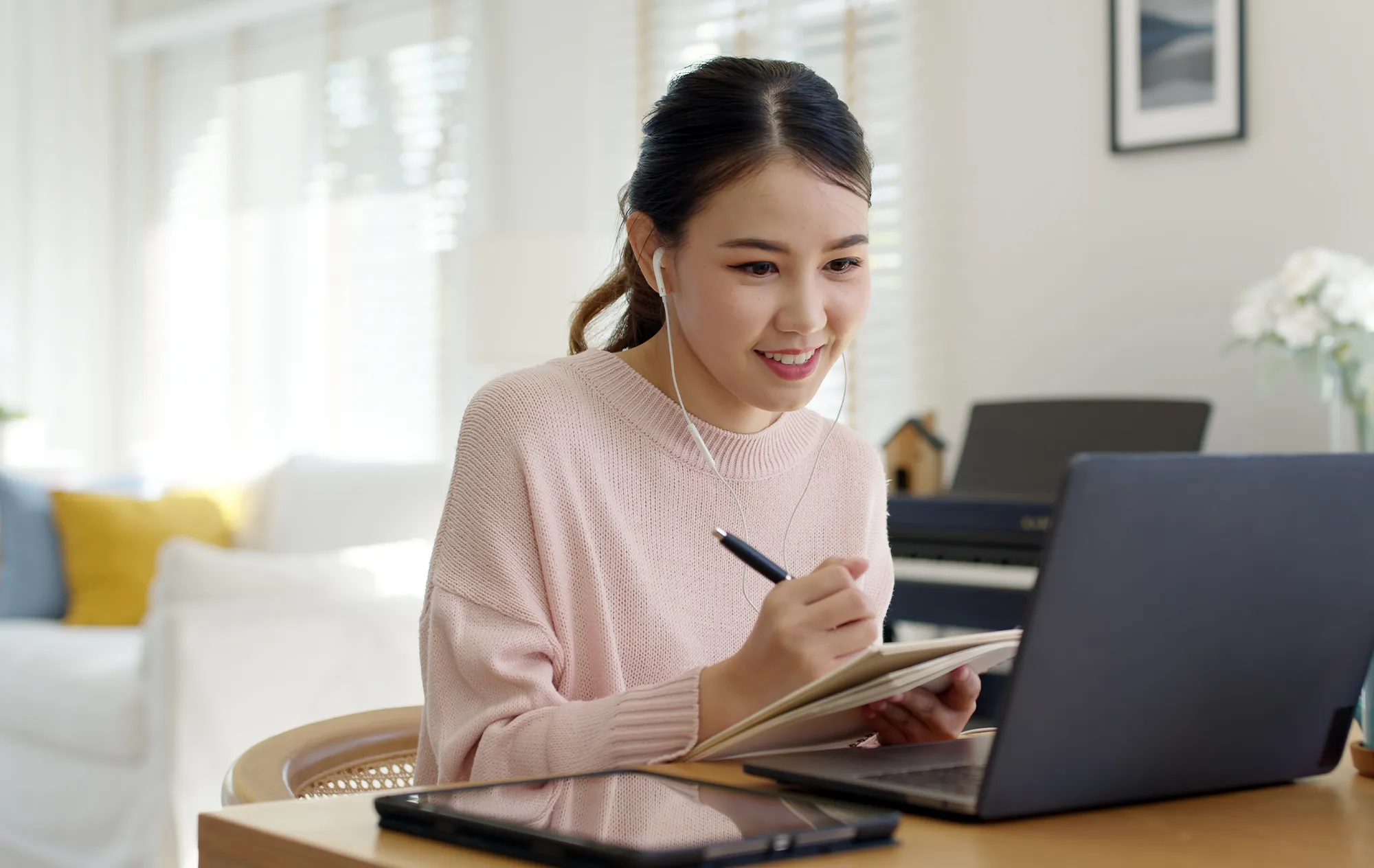 A smiling young woman in a pink jumper and white earphones writing in a notebook in front of a laptop, a tablet on the table