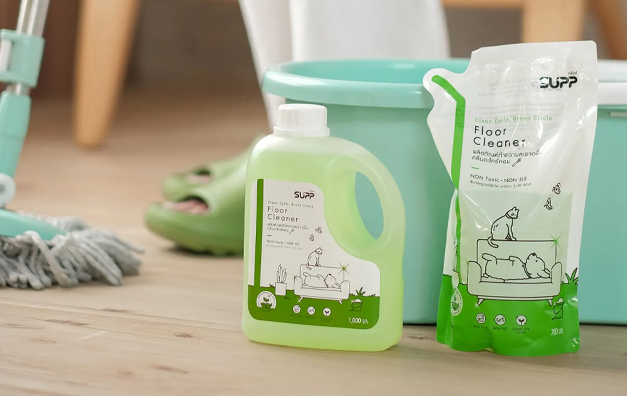 A bottle and a refill pouch of SUPP Floor Cleaner on a wooden floor beside a teal bucket, a mop and a pair of green slippers