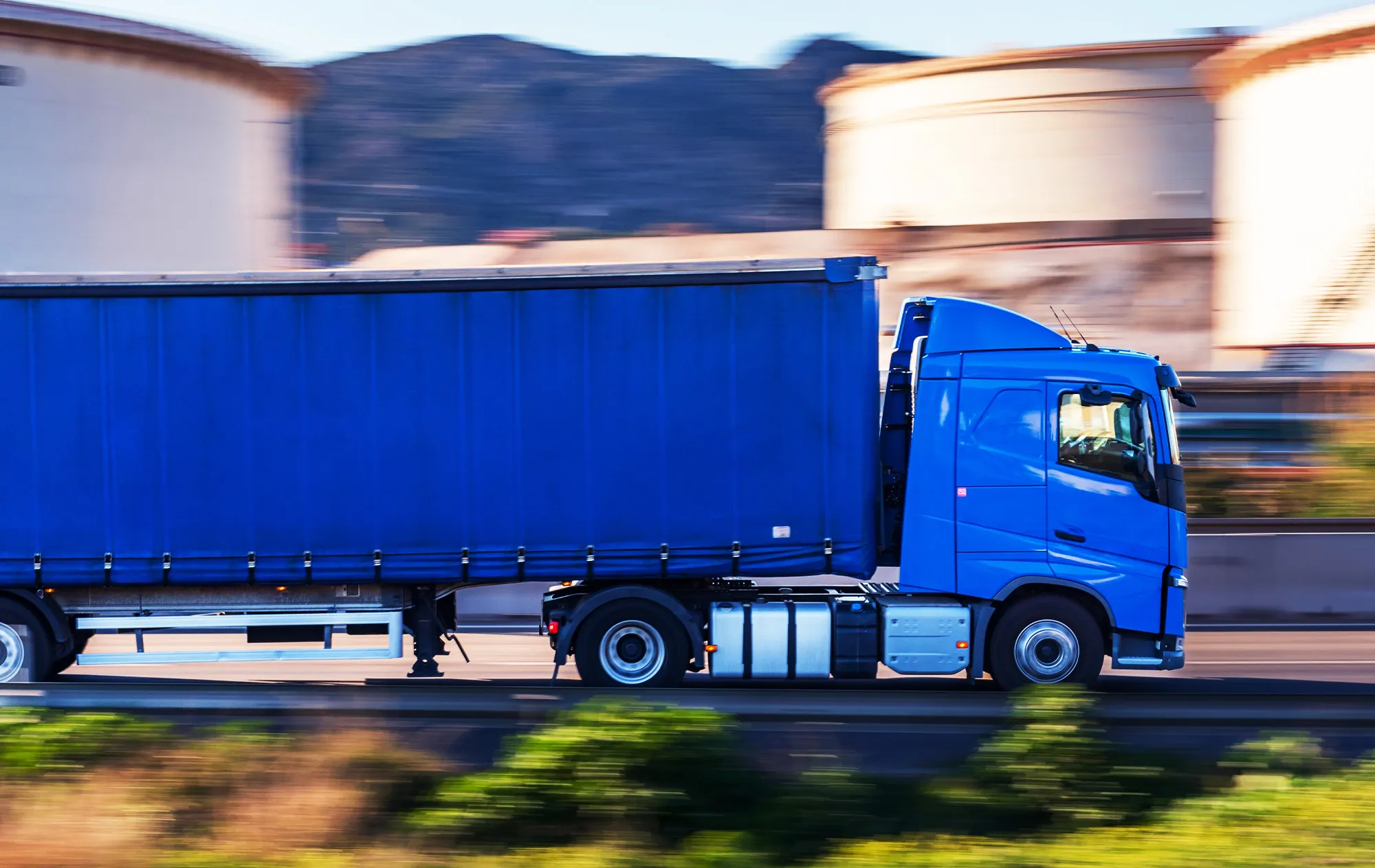 A blue truck and its blue curtain-sided trailer driving past large round storage tanks, the background blurred by motion