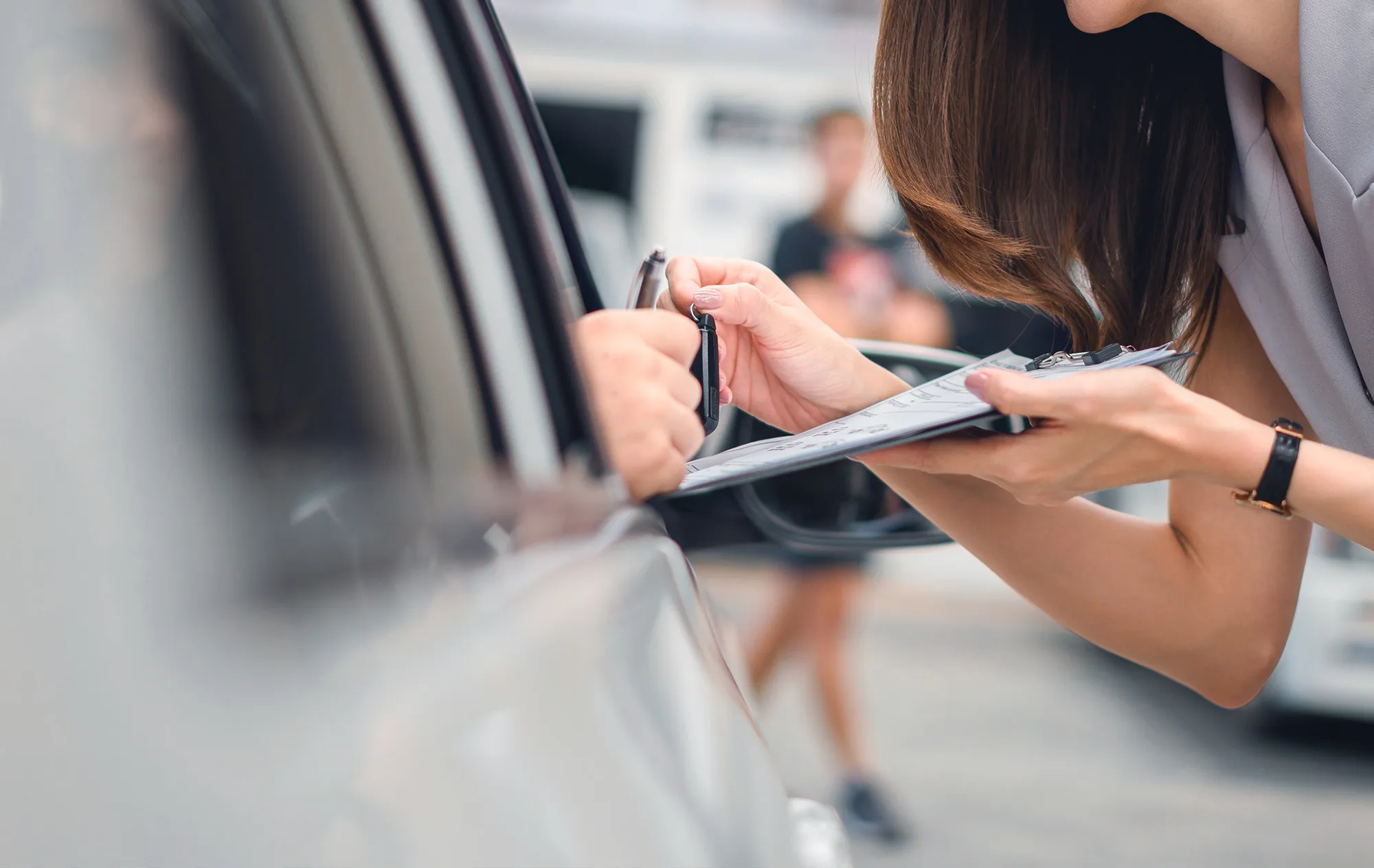A woman with long brown hair leaning towards a car window with a clipboard, her hand and the driver’s hand on a car key