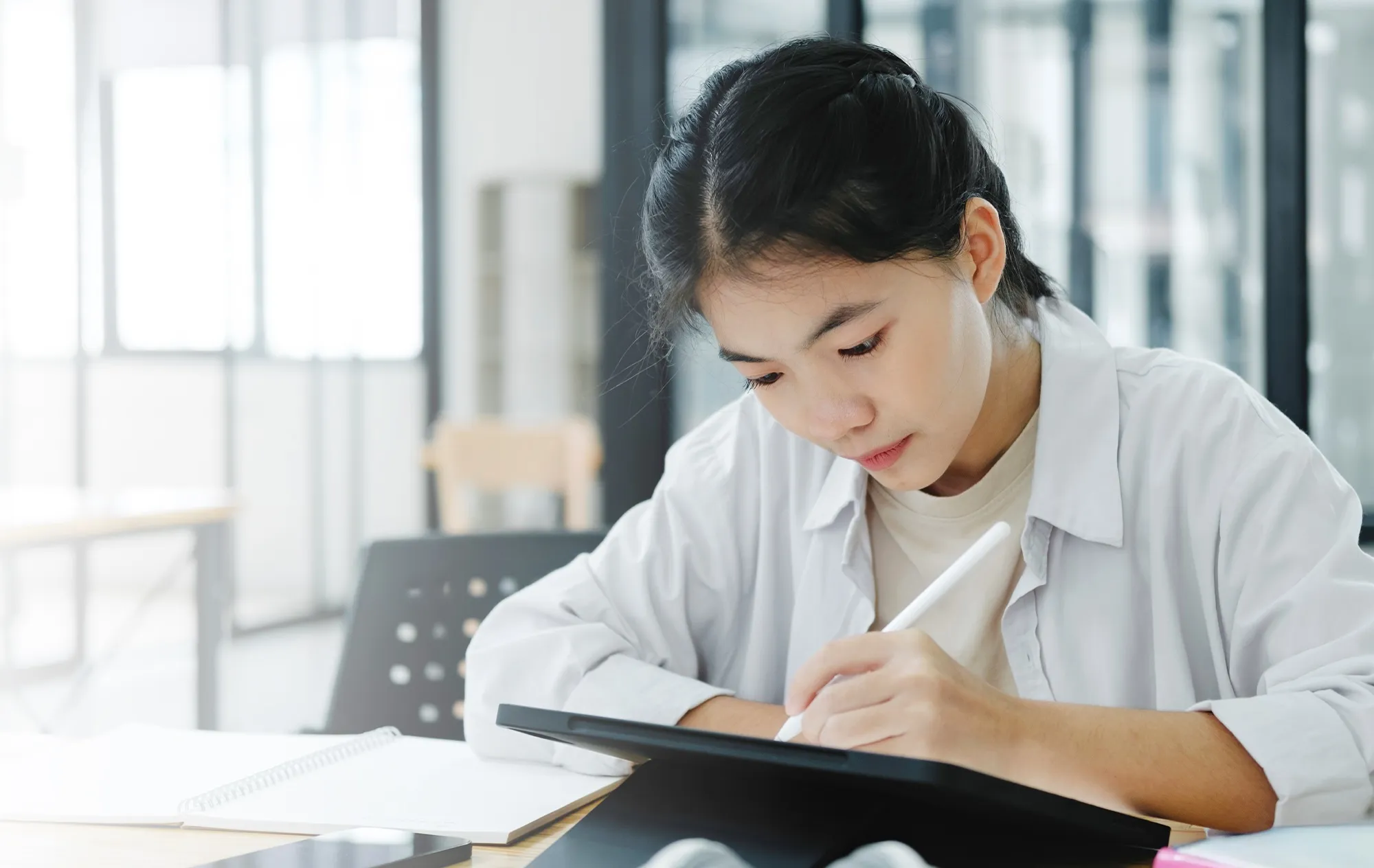 A young woman in a white shirt writing on a tablet with a stylus at a desk beside an open spiral notebook, tall windows behind her
