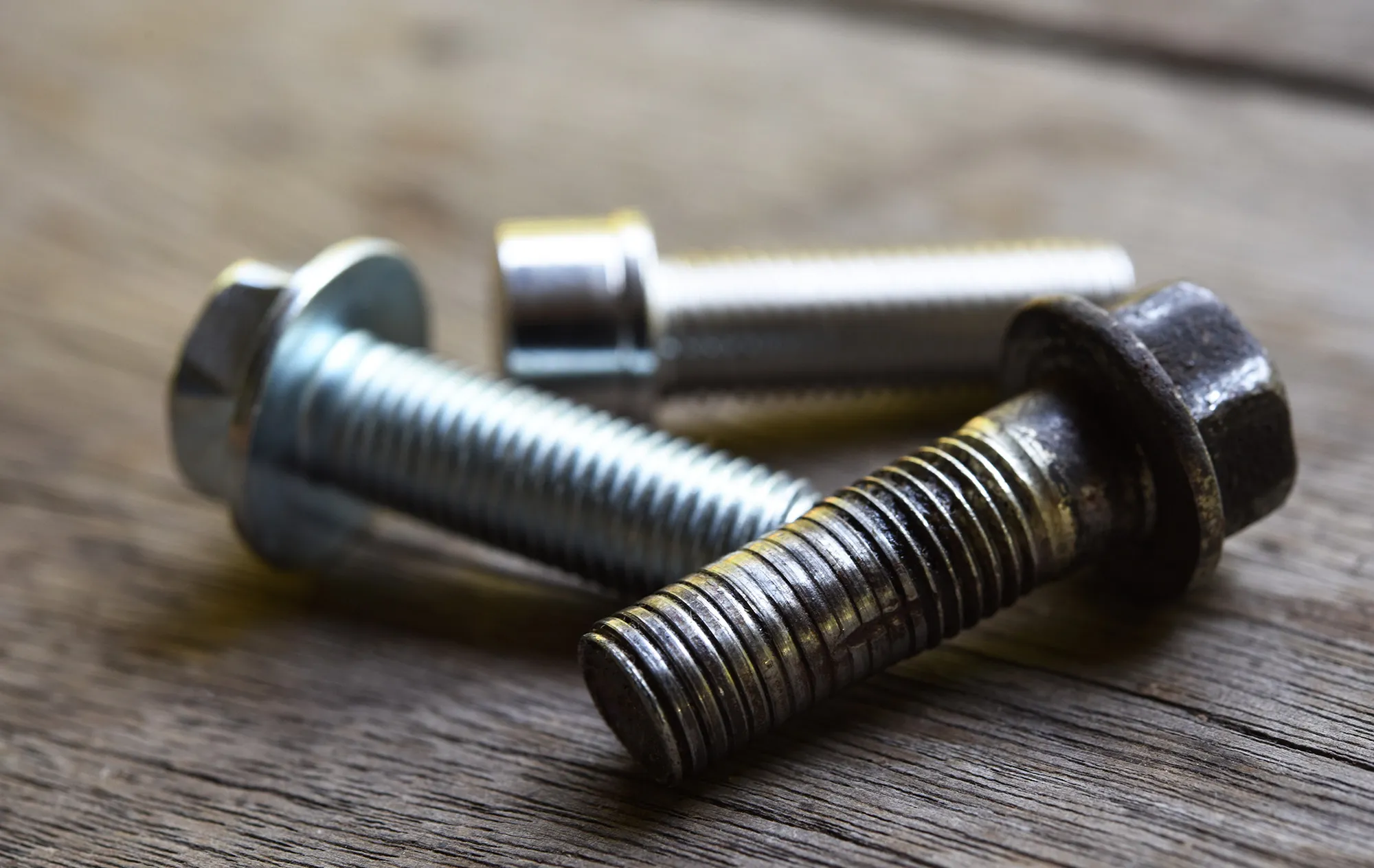 Three steel bolts lying on a weathered wooden table, a dark, worn bolt in front of two bright silver ones