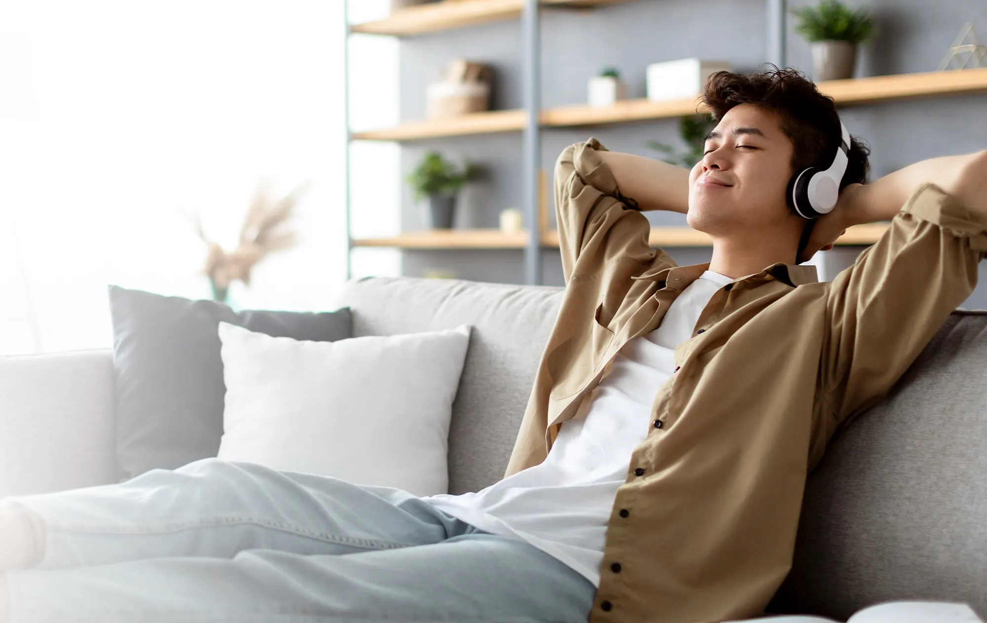 A young man in white headphones leaning back on a grey sofa with his hands behind his head and his eyes closed