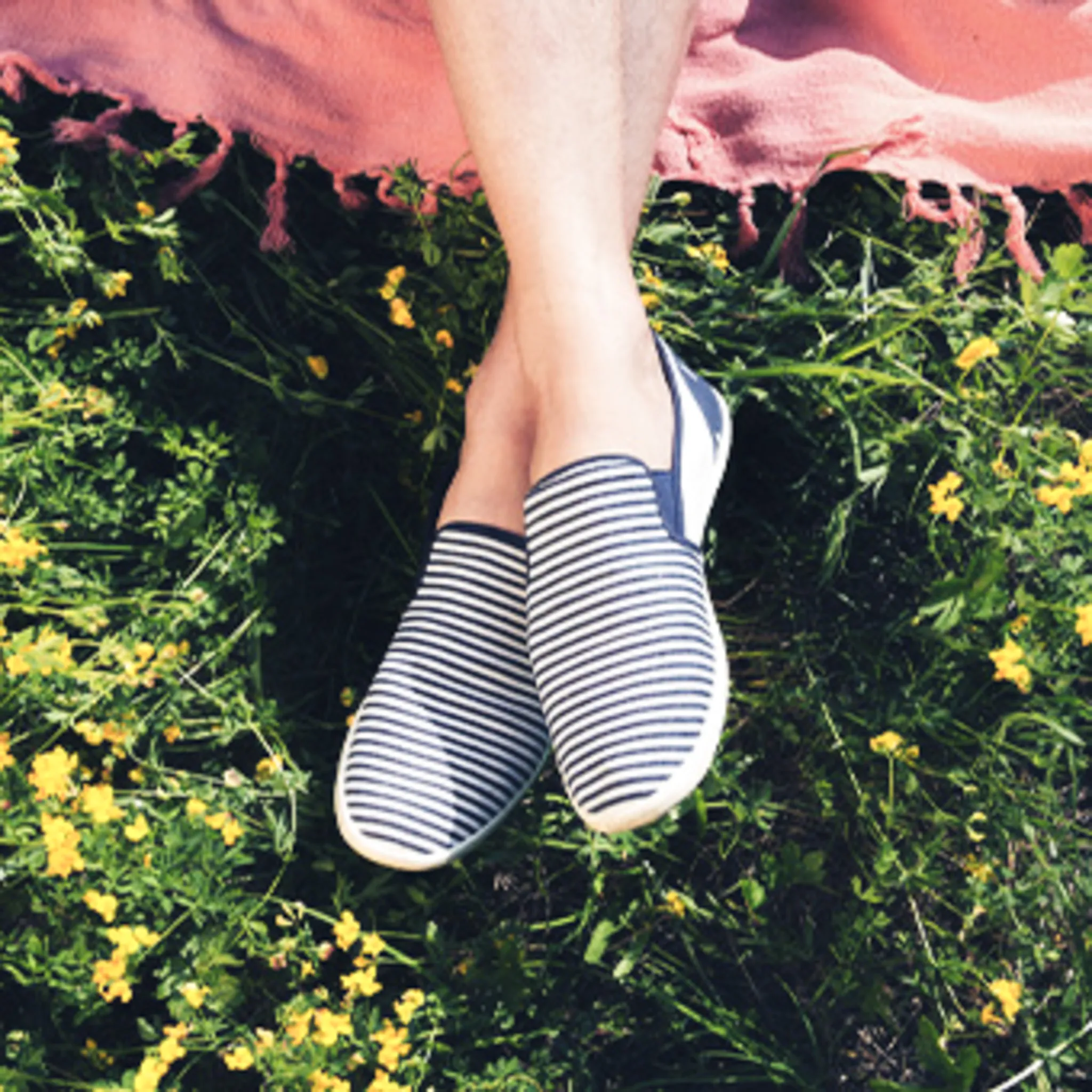 Legs crossed at the ankles in navy and white striped slip-on shoes, above grass with small yellow flowers and the fringed edge of a pink blanket