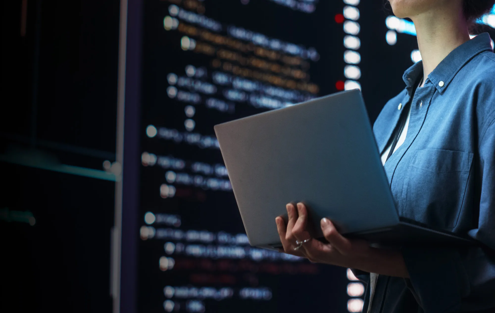 A woman in a blue denim shirt holding an open laptop in front of a large dark screen of blurred code