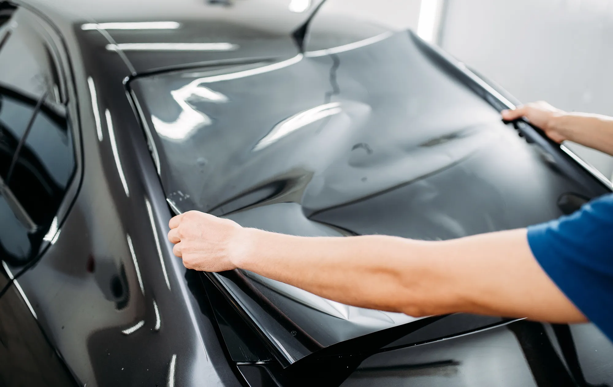 Two hands stretching a large sheet of dark tint film across the window of a black car