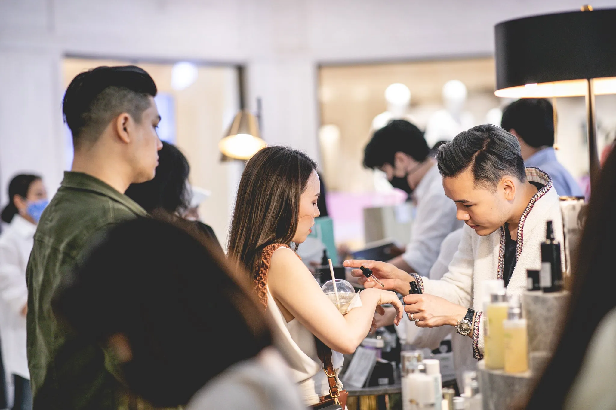 A man in a white tweed jacket holding a dropper over a woman’s wrist at a skincare counter, with other people around them