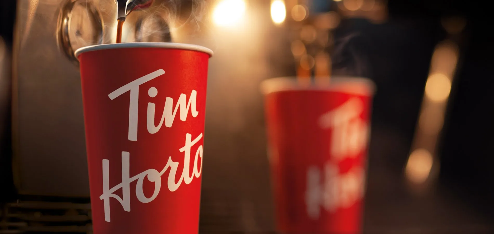 A red Tim Hortons cup with coffee pouring into it from a spout above and steam rising, with a second red cup out of focus behind it
