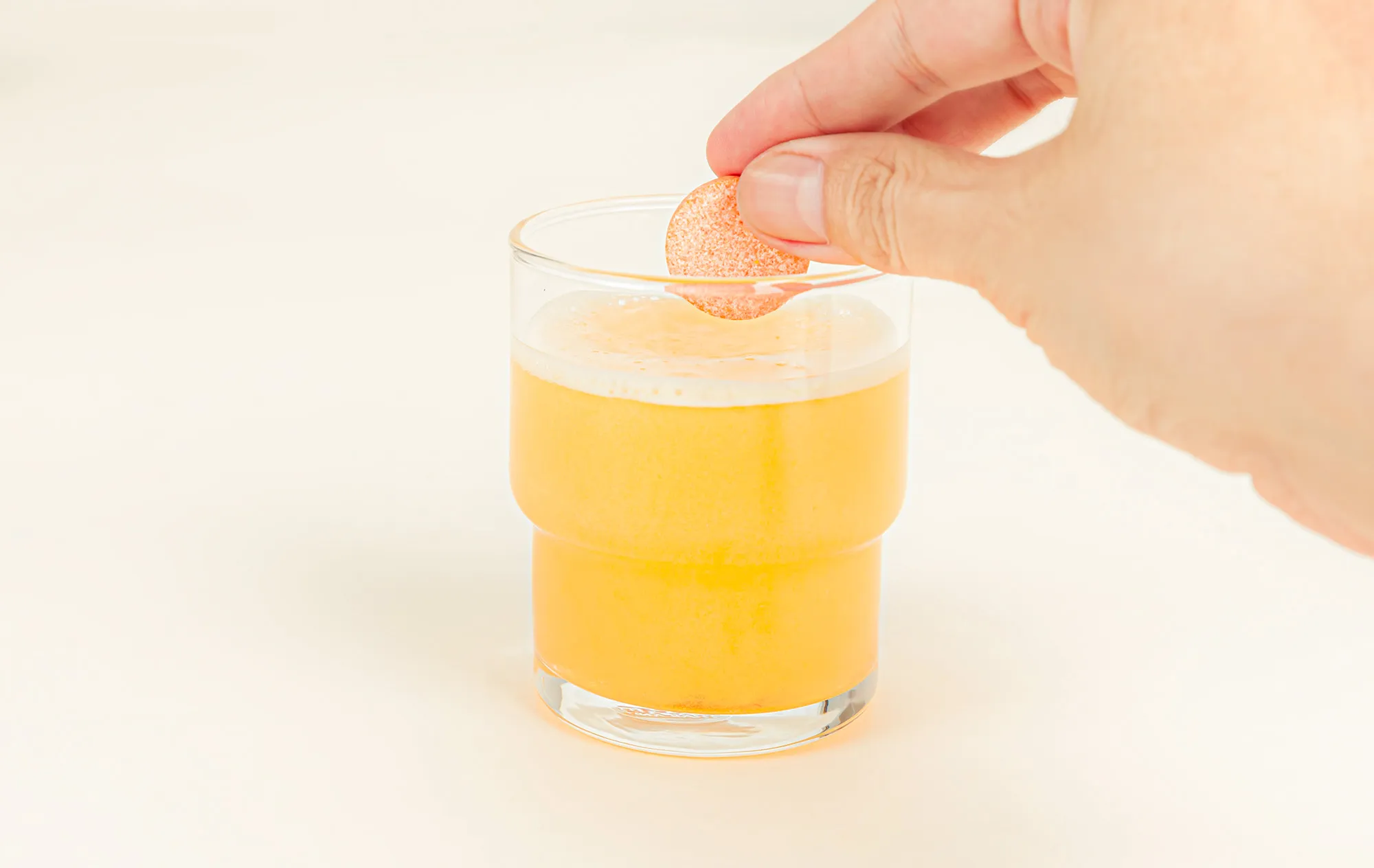 A hand dropping a round orange tablet into a glass of foaming yellow drink on a cream background