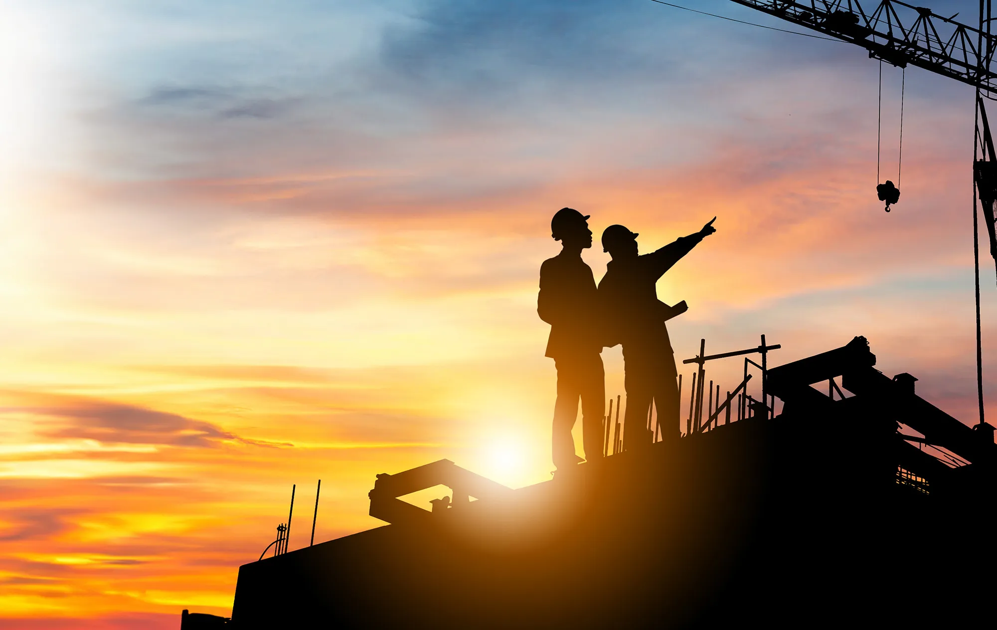 Two people in hard hats, seen in silhouette against an orange sunset sky, standing on a building under a crane hook, one pointing into the distance