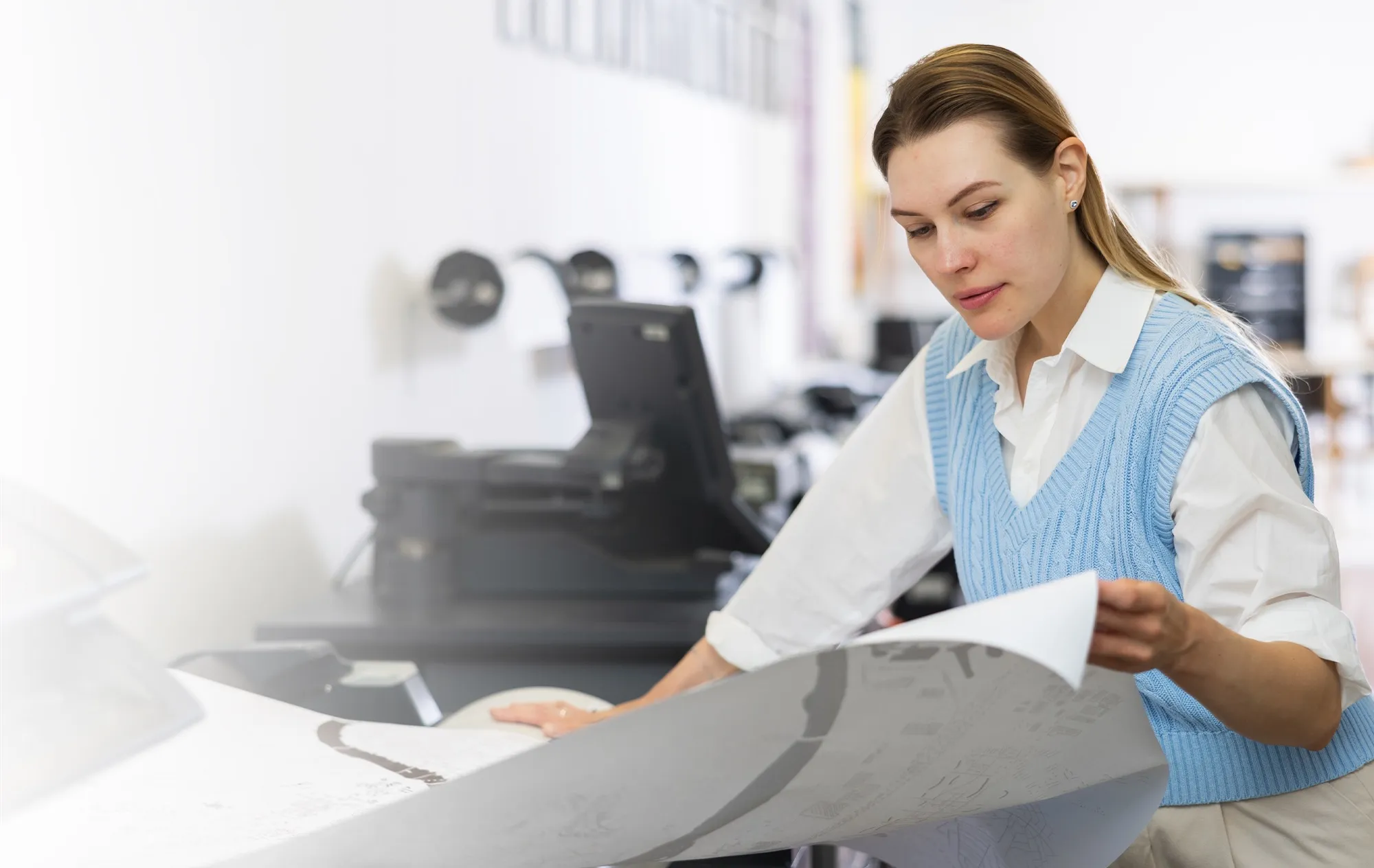 A woman in a white shirt and a blue knitted vest looking over a large printed map beside a wide-format printer