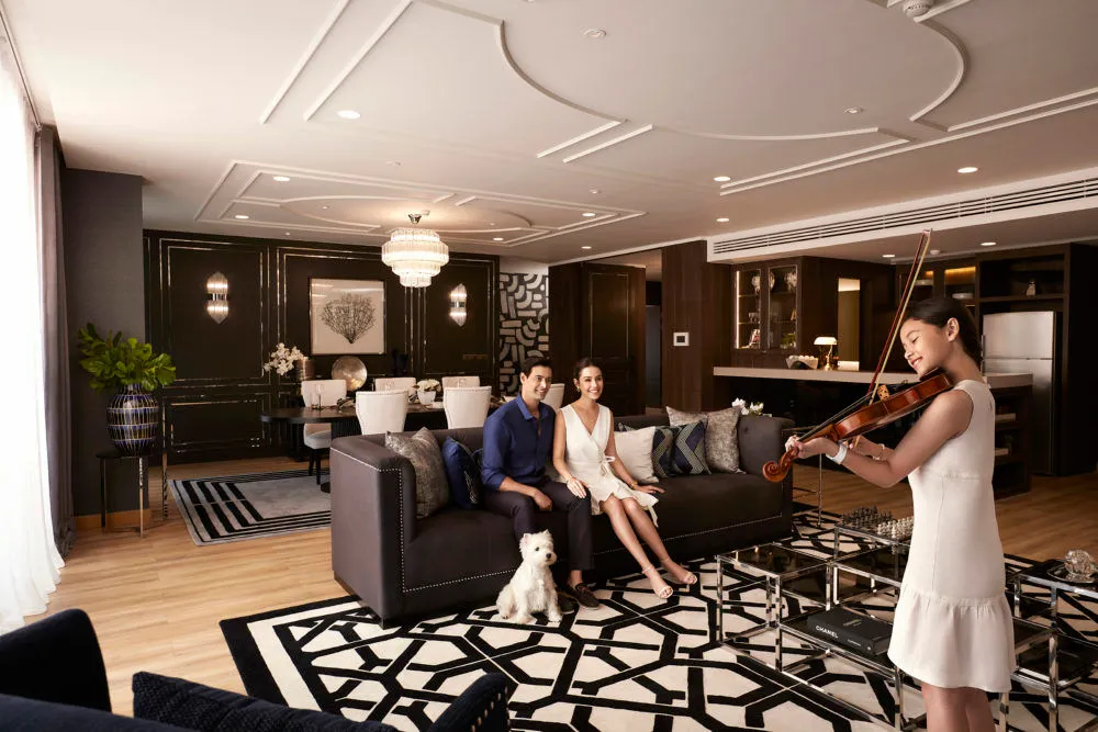 A couple on a dark sofa with a small white dog while a girl plays the violin in a large, modern lounge
