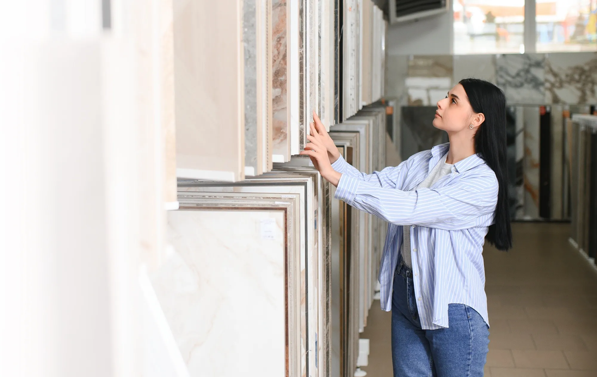 A woman in a striped shirt looking through large stone-effect tile samples on a display rack in a tile showroom