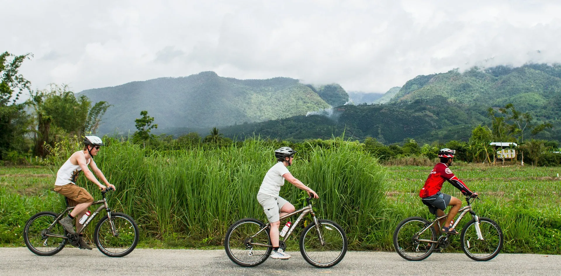 3 cyclists in helmets riding along a country road past tall green grass and fields, with forested mountains under low cloud behind them
