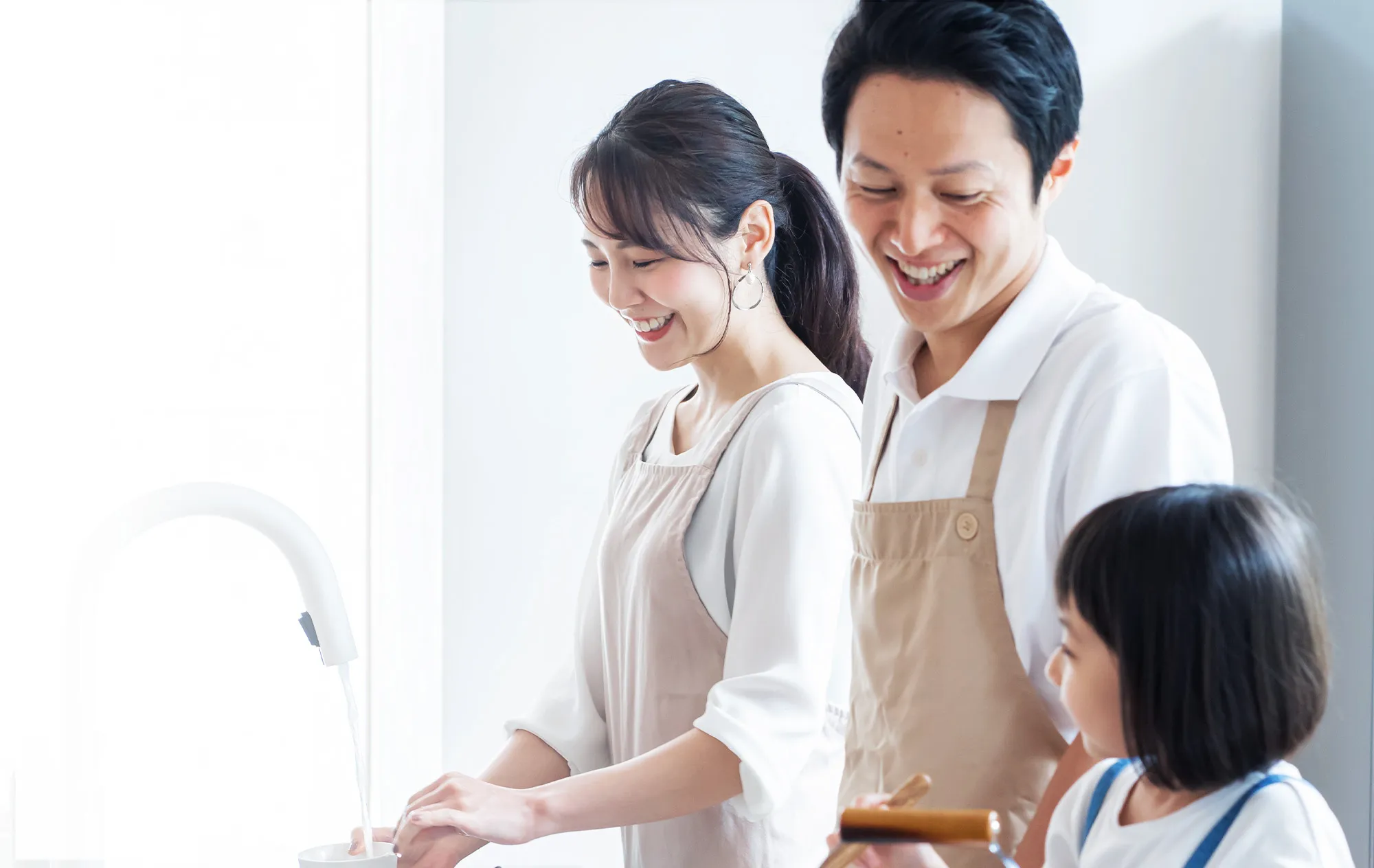 A smiling woman and man in beige aprons cooking at a white kitchen counter, the woman holding a cup under a running tap and a young girl beside them