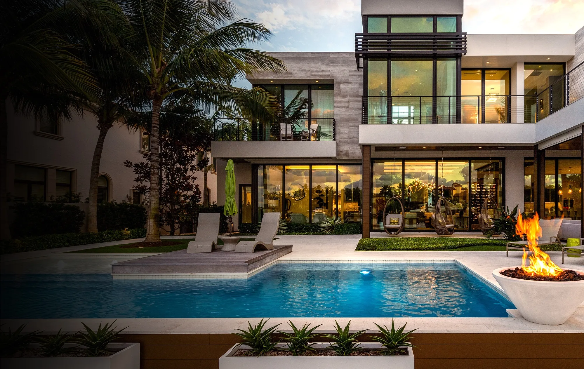A modern two-storey house with glass walls at dusk, with a pool, sun loungers, a fire bowl and palm trees
