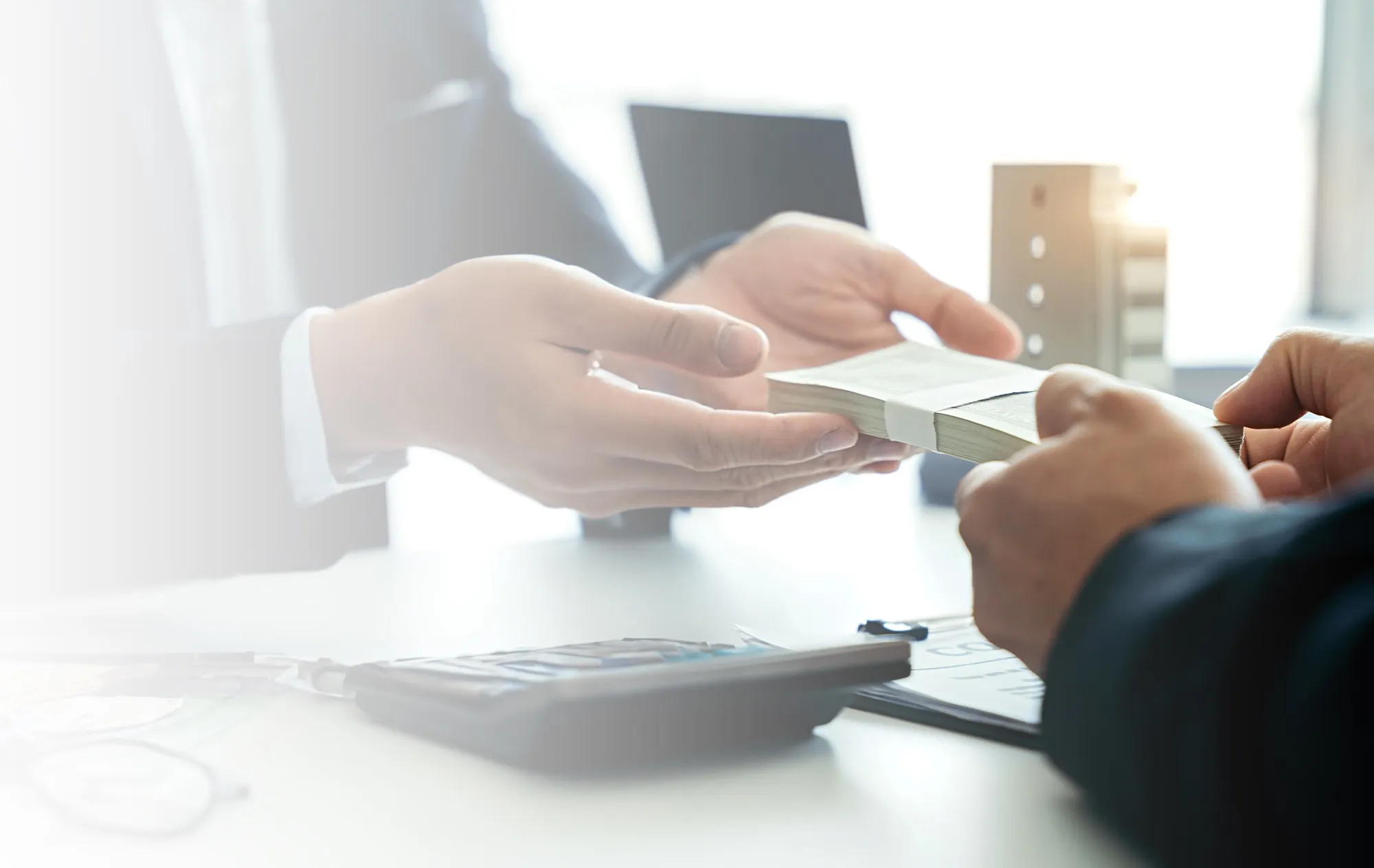 A hand in a dark sleeve passing a banded stack of banknotes to the open hands of a man in a suit across a white desk, a calculator in front