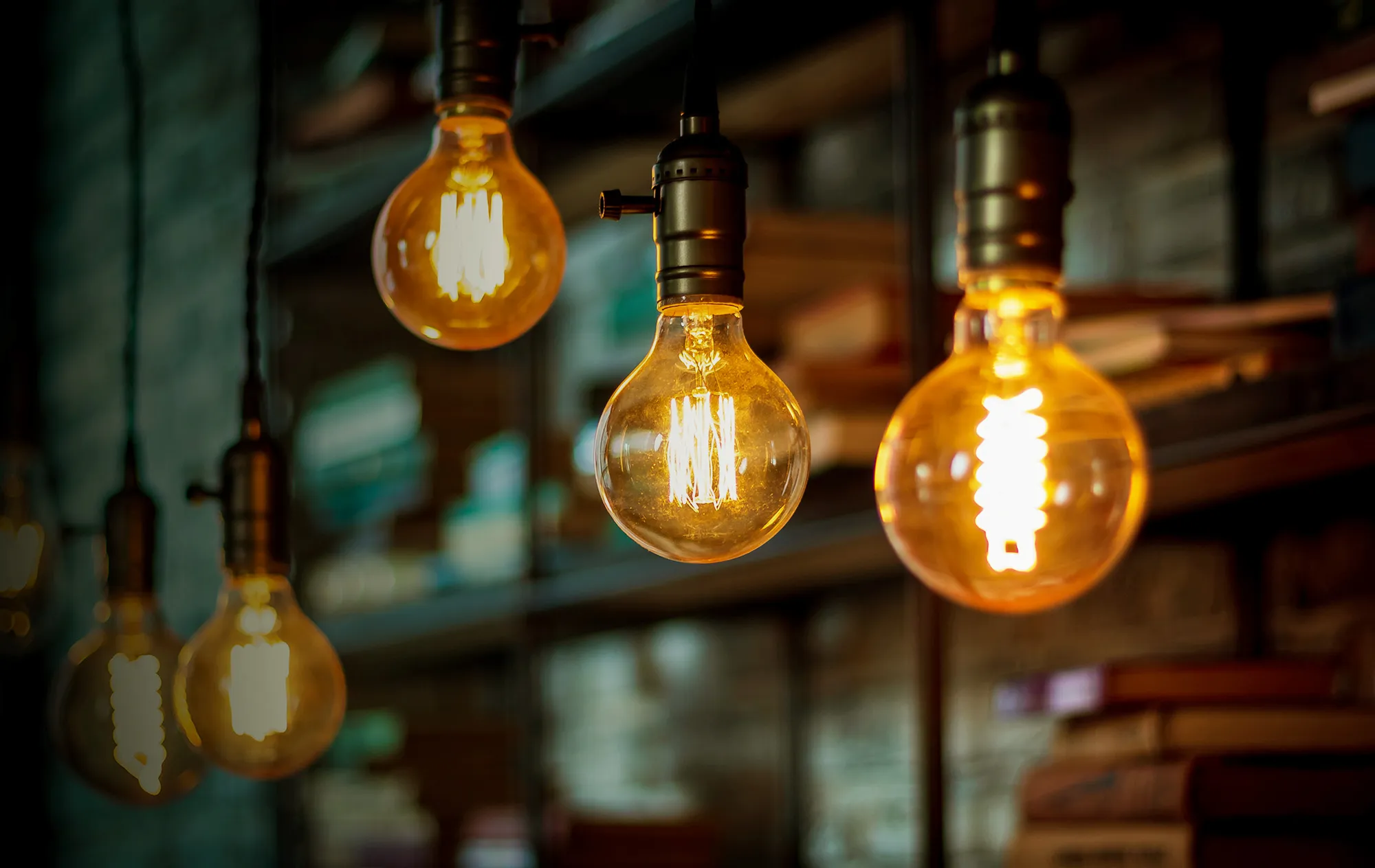 Round glass light bulbs with glowing filaments hanging from brass holders in front of blurred shelves