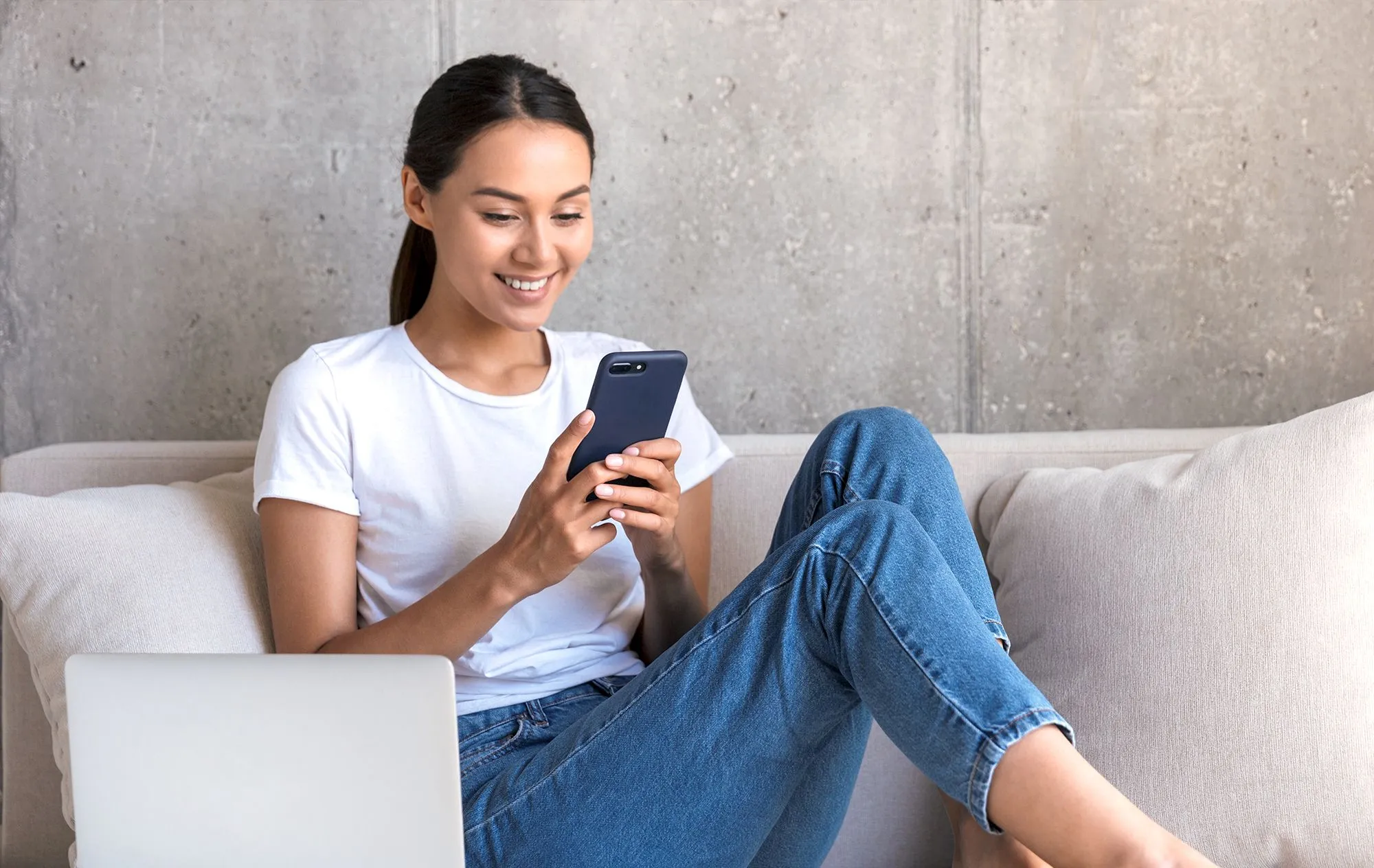 A smiling woman in a white T-shirt and jeans looking at her phone on a beige sofa, with a laptop in front of her and a concrete wall behind