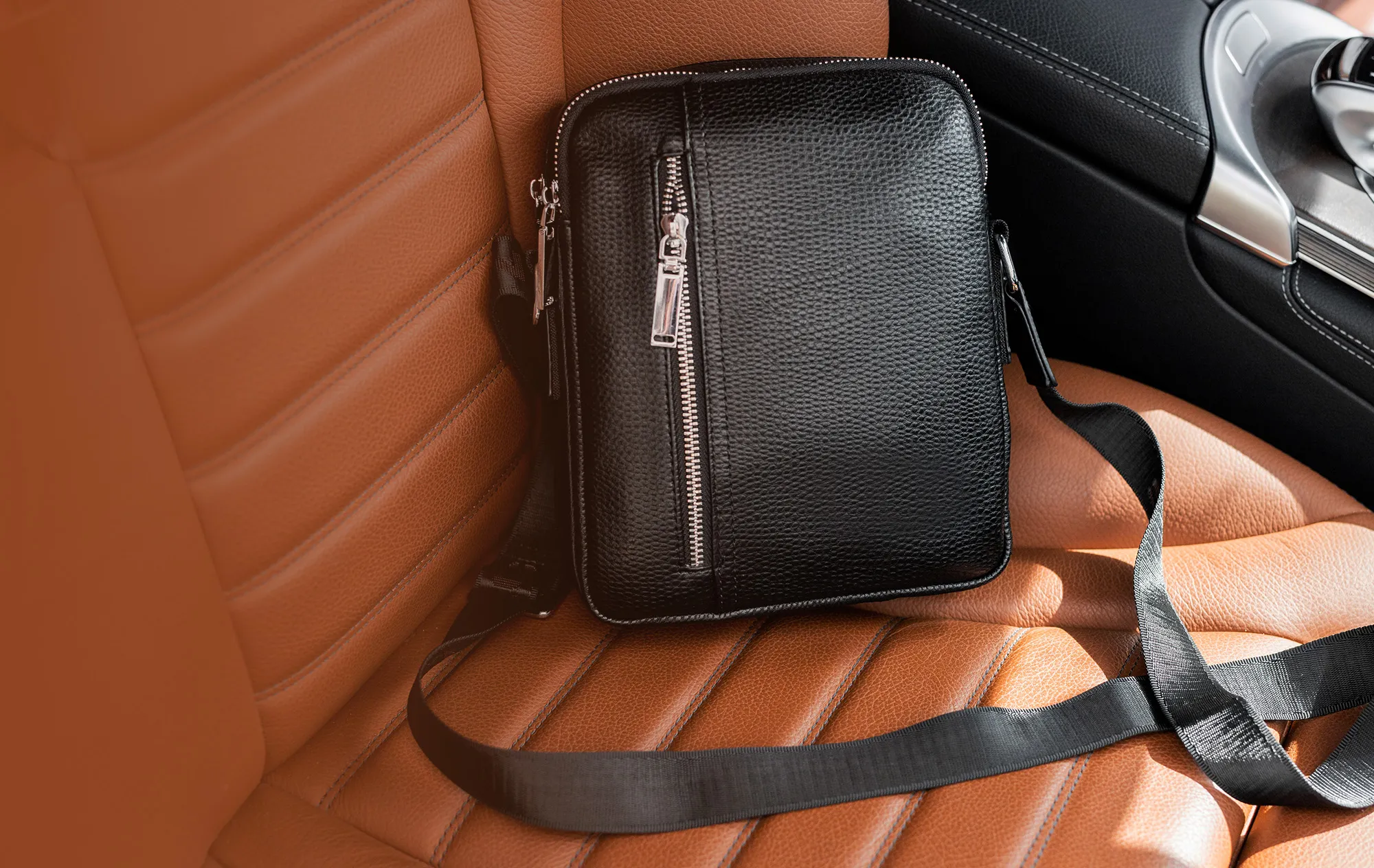 A black leather crossbody bag with a silver zip on the tan leather seat of a car