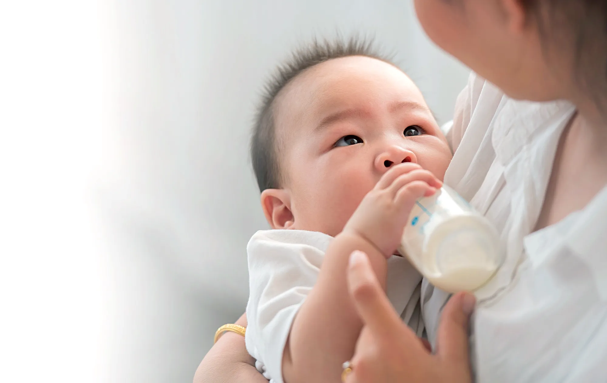 A baby in a white top drinking milk from a bottle in a woman’s arms, looking up at her