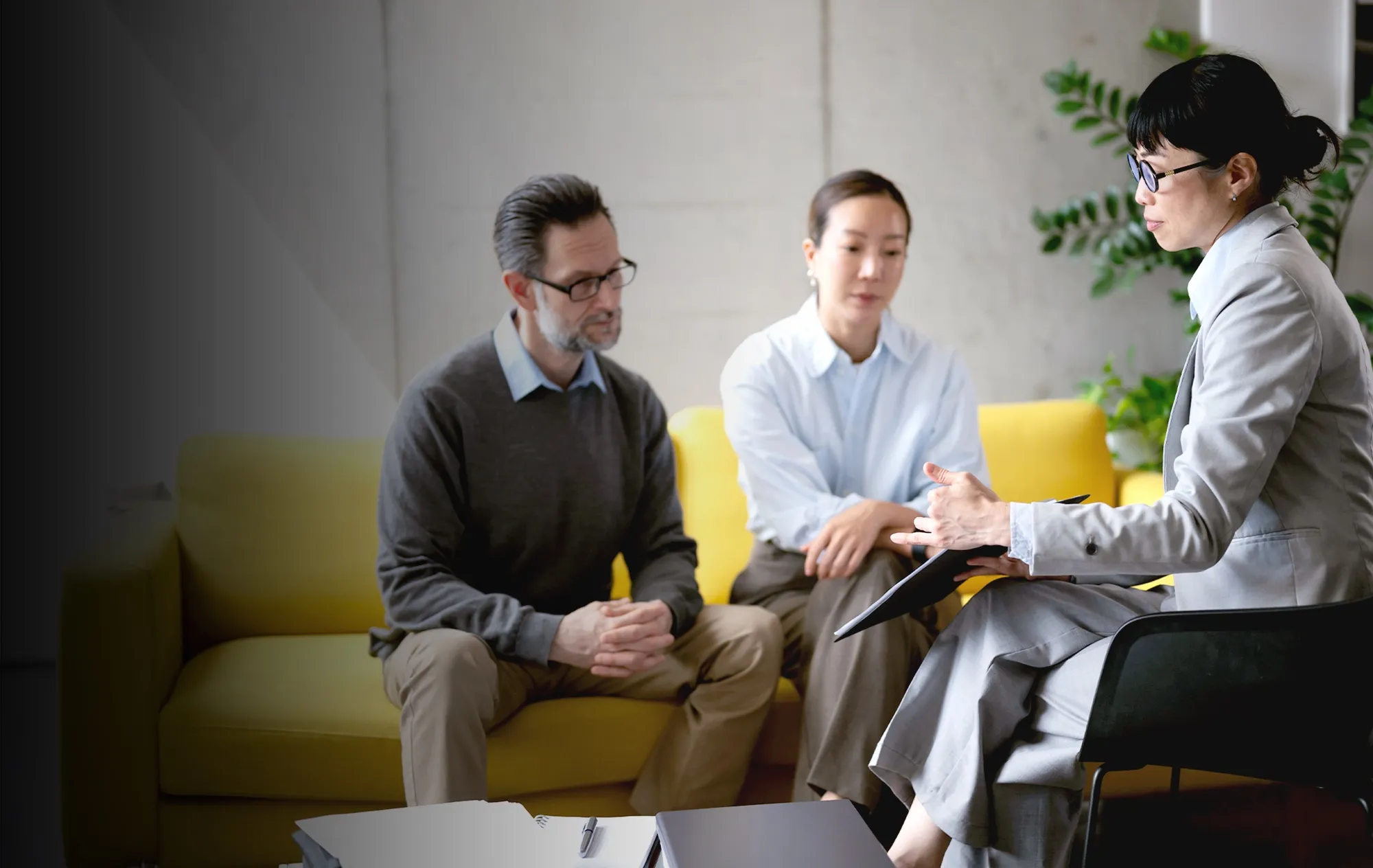 A woman in glasses and a grey suit holding a folder and talking with a man and a woman seated on a yellow sofa
