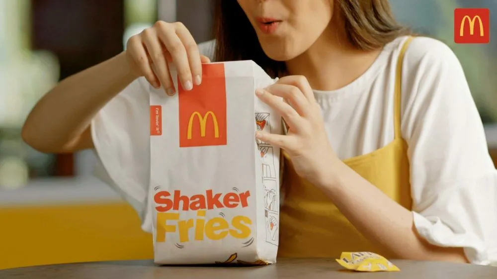 ผู้หญิงถือถุง Shaker Fries ของ McDonald’s มีซองผงวางอยู่บนโต๊ะตรงหน้า