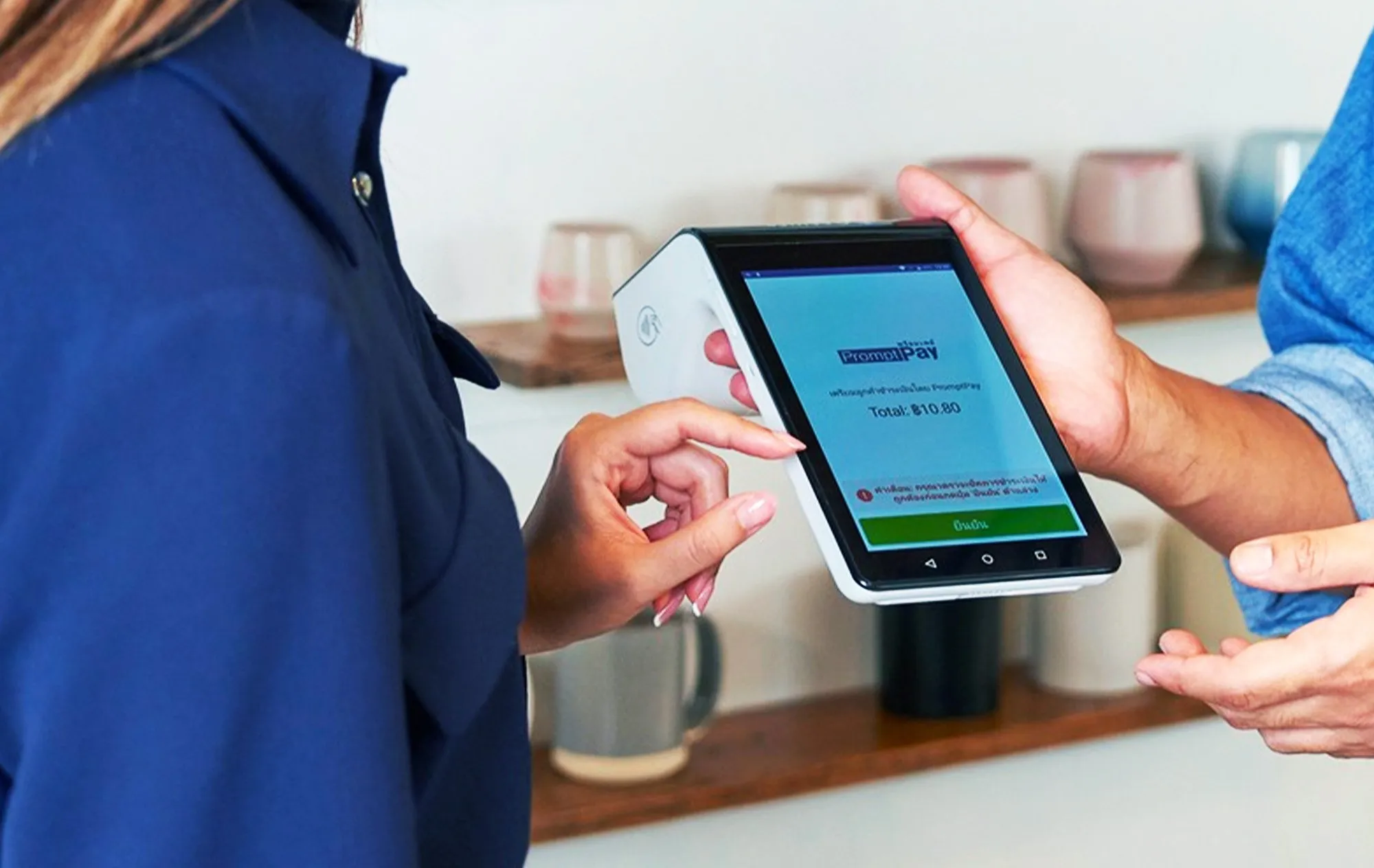 A payment terminal showing a PromptPay screen with a total, with one hand holding its top edge and another hand pointing at the screen