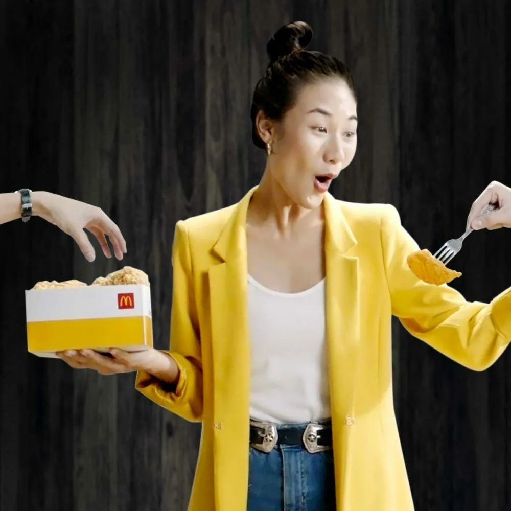 ผู้หญิงสวมเสื้อสูทสีเหลืองถือกล่องไก่ของ McDonald’s มองไก่บนส้อมที่ยื่นมาให้อย่างประหลาดใจ