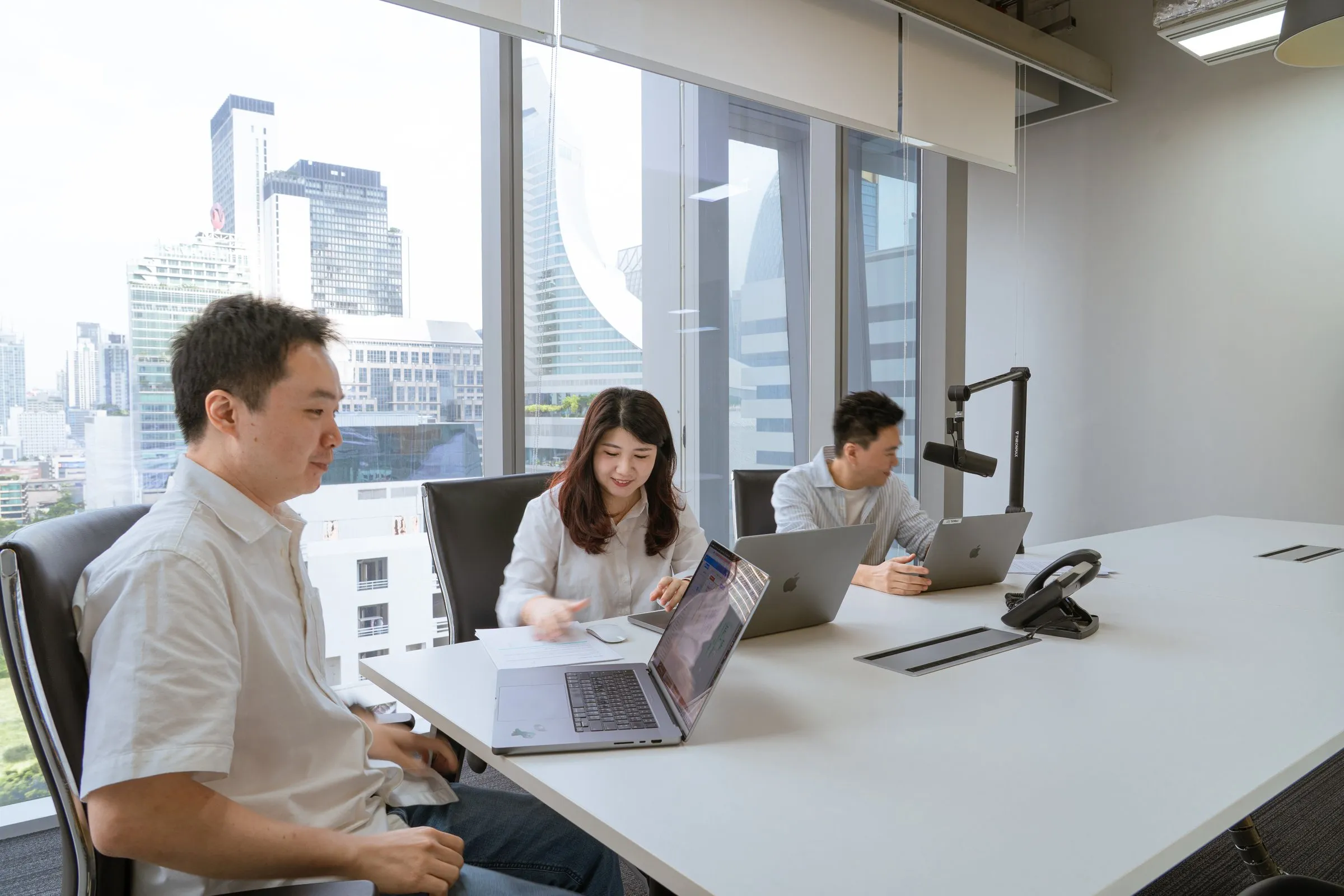 Three colleagues working on laptops at a long white meeting table beside tall windows with a view over the city