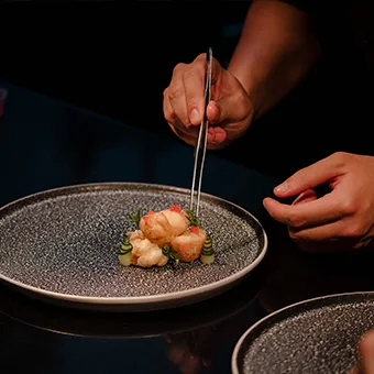 Hands using tweezers to place prawns and cucumber on a dark speckled plate