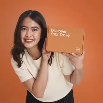 ผู้หญิงยิ้มชูกล่องสีส้มที่พิมพ์คำว่า Discover Your Smile และสัญลักษณ์ของ SELFIE