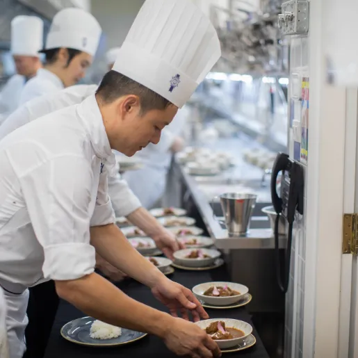 A chef in a tall white hat setting bowls of food in a row along a counter, with other chefs at work behind him
