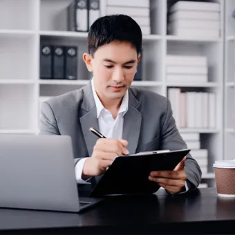 A man in a grey suit writing on a clipboard at a desk beside a laptop and a paper cup, shelves of files behind him