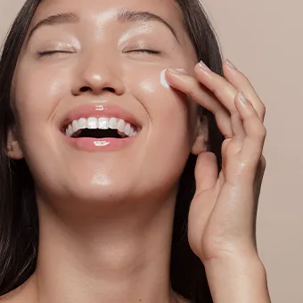 A woman laughing with her eyes closed as she spreads white cream across her cheek
