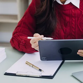 A woman in a red cable-knit jumper holding a white stylus over a tablet, with a clipboard and a gold pen on the desk