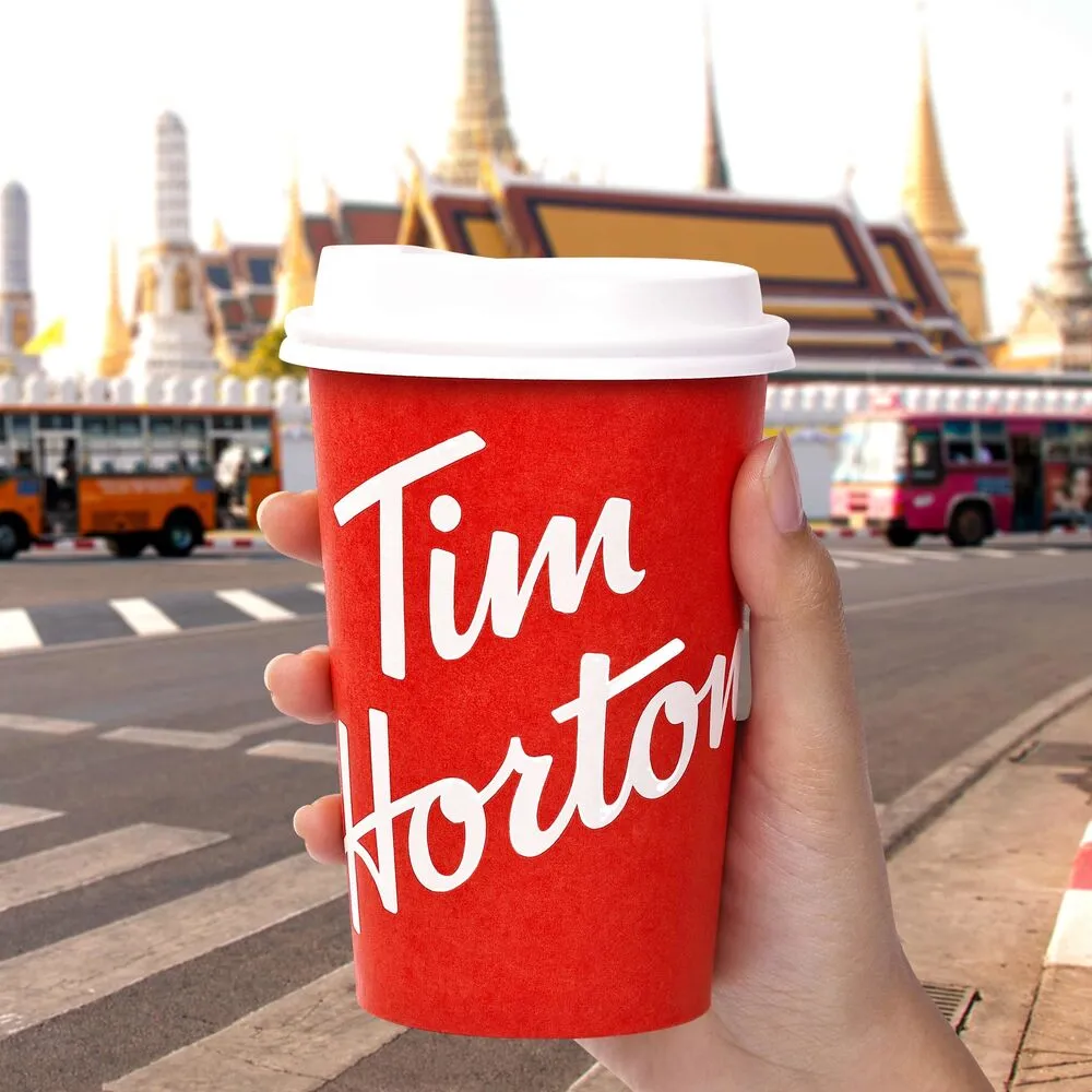 มือถือแก้วกาแฟสีแดงของ Tim Hortons บนถนนในกรุงเทพฯ ด้านหลังมียอดวัดและรถเมล์
