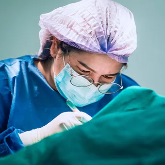 A doctor in a surgical cap, face mask and glasses working over green surgical drapes
