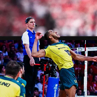 A volleyball player in a yellow shirt drawing back his arm to hit the ball at the net while a referee with a headset watches from behind