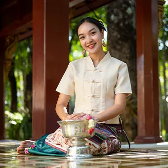 A smiling woman in a cream Thai-style blouse and a patterned skirt sitting beside a silver bowl of flowers, between wooden pillars