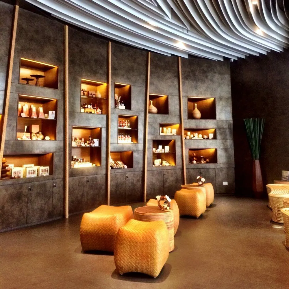 Lit shelves of spa products and vases set into a dark stone wall, with woven rattan stools in front