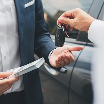A hand passing a car key to a man in a suit who is holding a stack of banknotes