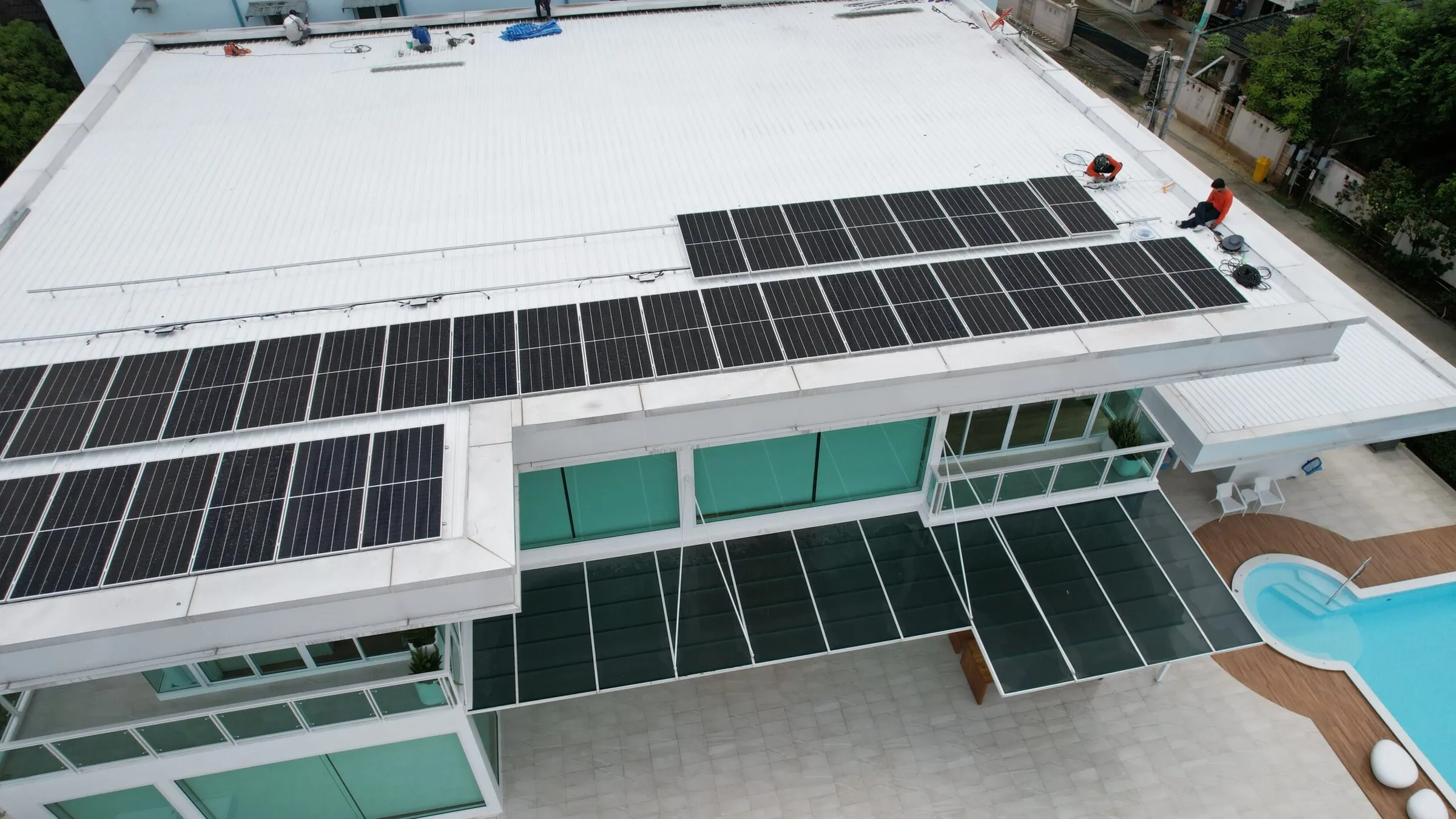 Aerial view of rows of solar panels on a white roof, with 2 workers in orange tops beside the panels and a glass canopy below