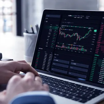Hands typing on a laptop that shows candlestick charts and price tables on a dark screen